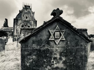 Nahaufnahme eines alten, verwitterten Grabsteins auf dem Friedhof der Sweetheart Abbey; oben thront ein Relief mit Davidstern, im Hintergrund ist die unscharfe Silhouette der Kirchenruine zu erkennen.