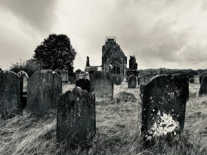 Perspektivische Ansicht mehrerer dunkler, abgerundeter Grabsteine, die in Richtung der massiven, eingerüsteten Ruine der Sweetheart Abbey führen.