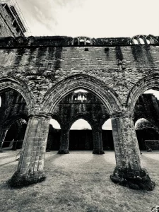 Schwarzweißaufnahme der inneren Klosterruine der Sweetheart Abbey mit massiven steinernen Bögen und Säulen, die einen offenen, leeren Innenraum umrahmen.