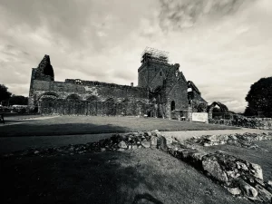 Großaufnahme der langen Seitenwand der Sweetheart Abbey mit einer Reihe von blinden Arkaden und den hohen, eingerüsteten Türmen unter einem dramatischen Wolkenhimmel.