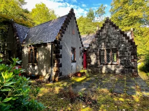 Historisches Abbot’s House in New Abbey, Schottland: Ein steinernes Gebäude mit charakteristischen Treppengiebeln, grauer Fassade und einer auffällig leuchtend roten Tür, umgeben von grünem Garten unter leicht bewölktem blauem Himmel.