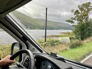 Sicht aus der Windschutzscheibe eines Fahrzeugs. Man sieht die Hand des Fahrers am Lenkrad und den Tacho, der 45 mph anzeigt. Draußen zieht eine regnerische Seenlandschaft mit Bergen vorbei.