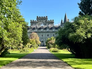 Ein imposantes, graues Schloss mit spitzen Türmen am Ende eines geraden Kieswegs. Der Weg ist von grünen Rasenflächen, Bäumen und gepflegten Sträuchern gesäumt.