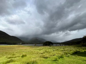 Weite Landschaftsaufnahme eines grünen Feldes im Vordergrund, das zu einem See führt. Im Hintergrund stehen die steinernen Ruinen einer Burg vor dunklen, dramatischen Regenwolken und Bergen.