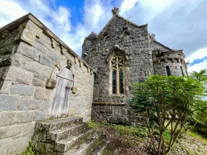 Eine alte Holztür in einem steinernen Anbau der Kirche, erreichbar über grobe Steinstufen, flankiert von grünen Sträuchern.