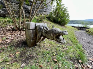 Eine hölzerne Skulptur eines Drachenkopfes, die wie ein Wasserspeier aus einer Steinmauer ragt, im Hintergrund der See.