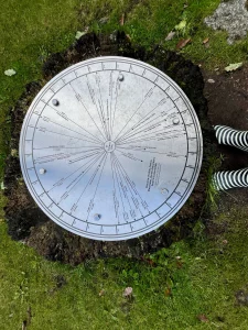 Eine große Sonnenuhr aus Metall im Vordergrund mit einem malerischen Blick auf einen weiten See und Hügel im Hintergrund.