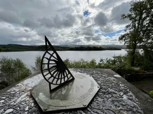 Eine runde Metallplatte auf einem Baumstumpf, die wie ein Kompass gestaltet ist und Entfernungen zu Städten und Bergen von St Conan’s Kirk aus anzeigt.