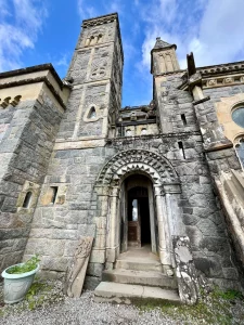 Frontalansicht eines hohen Steinturms und eines Rundbogen-Eingangs mit tiefen Steinmetzarbeiten; davor lehnen dekorative Steinplatten.