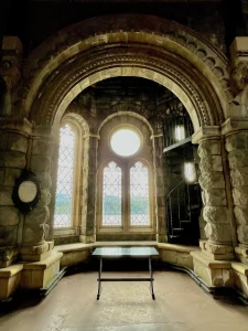 Ein massiver Steinbogen rahmt drei Fenster ein, durch die helles Licht fällt. Rechts ist eine filigrane Wendeltreppe aus Metall zu sehen.