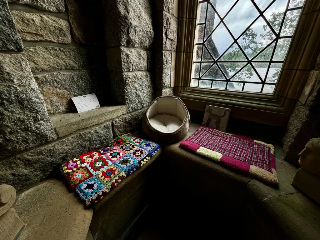 Ein gemütlicher Platz in einer tiefen Steinnische mit bunten Häkeldecken, einem kleinen Katzenbett und einem Blick nach draußen.