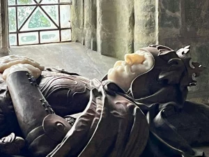 Seitenansicht des Gesichts und des Oberkörpers einer steinernen Grabfigur, die einen König in Rüstung mit Krone und gefalteten Händen darstellt.