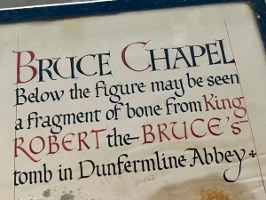Nahaufnahme einer handgeschriebenen Tafel in Schwarz und Rot. Der Text besagt, dass sich unter der Figur ein Knochenfragment aus dem Grab von König Robert the Bruce in der Dunfermline Abbey befindet.