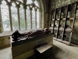 Innenansicht einer kleinen Steinkapelle mit einem großen Hochgrab, auf dem eine liegende Ritterfigur ruht. Ein großes Fenster mit Buntglas lässt helles Licht herein.