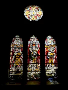 Drei schmale, spitzzulaufende Buntglasfenster zeigen Ritterfiguren; darüber ein rundes Fenster mit Engelsmotiven vor dunklem Hintergrund.