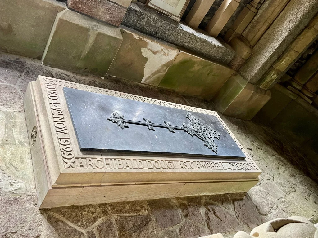 Ein rechteckiger Steinsarkophag an einer Bruchsteinwand, dessen Kanten mit Namen und Lebensdaten beschriftet sind und der oben eine dunkle Metallplatte mit einem verzierten Kreuz trägt.