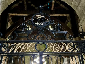 Blick nach oben auf ein schwarzes Eisengitter. Im oberen Bogen ist ein kreisförmiges Emblem mit einer goldenen Galeere integriert. Darunter befinden sich filigrane, goldfarbene Buchstaben und ein zentrales Herz-Symbol.