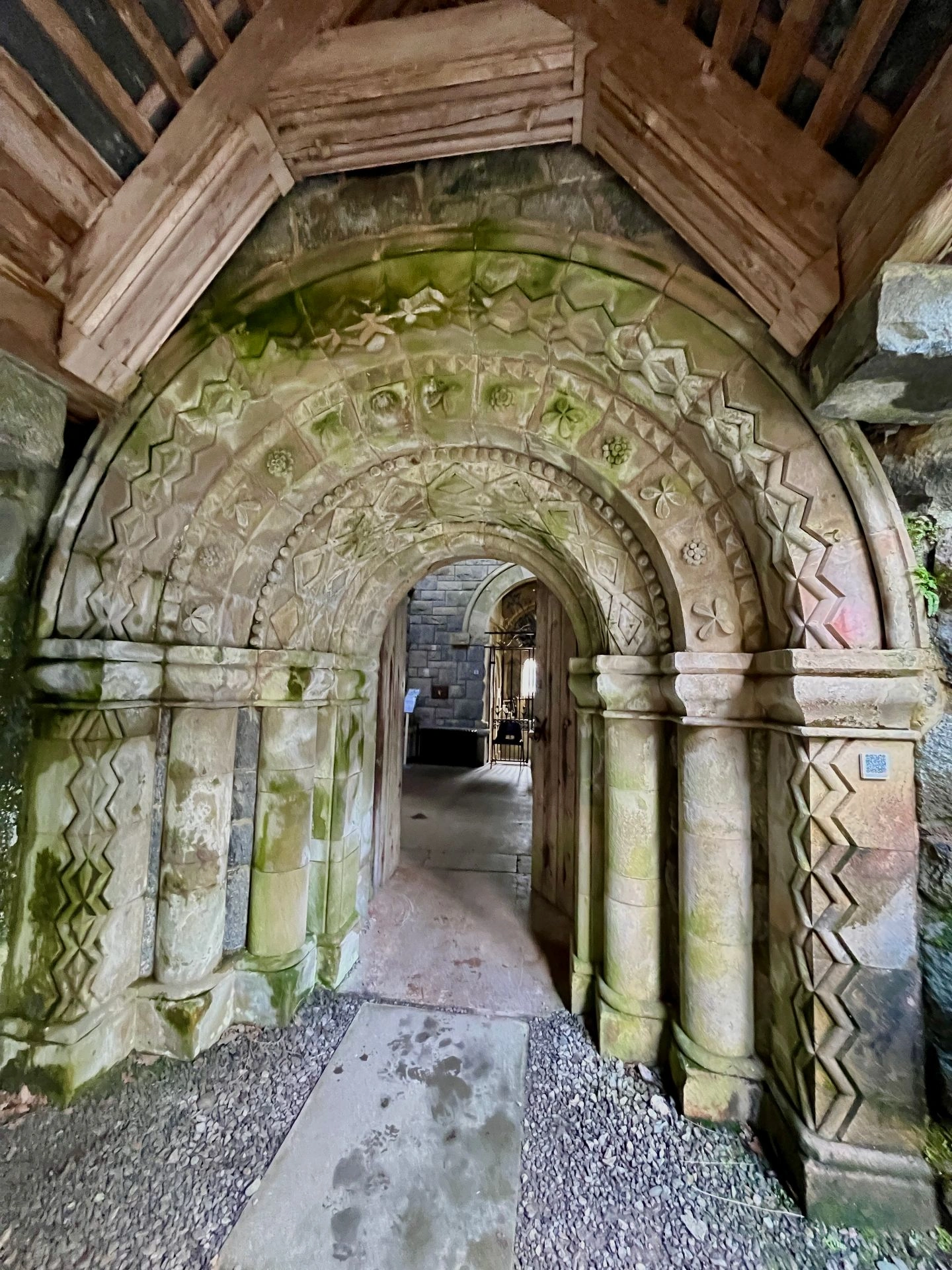 Frontale Ansicht eines tief gestaffelten Steinportals. Die massiven Rundbögen sind mit abwechslungsreichen geometrischen Mustern, Zickzack-Linien und floralen Reliefs verziert. Moosbewuchs verleiht dem hellen Stein eine grünliche Patina, während der Blick durch die offene Tür in den dunkleren Innenraum fällt.