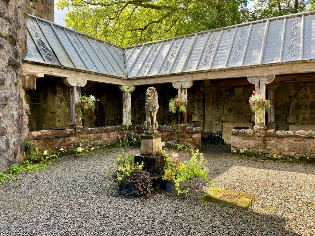 Blick in einen kiesbedeckten Innenhof der St. Conan’s Kirk. In der Mitte steht eine steinerne Löwenstatue auf einem Sockel, umgeben von Blumentöpfen. Ein hölzerner Kreuzgang mit Bleidach umschließt den Hof.