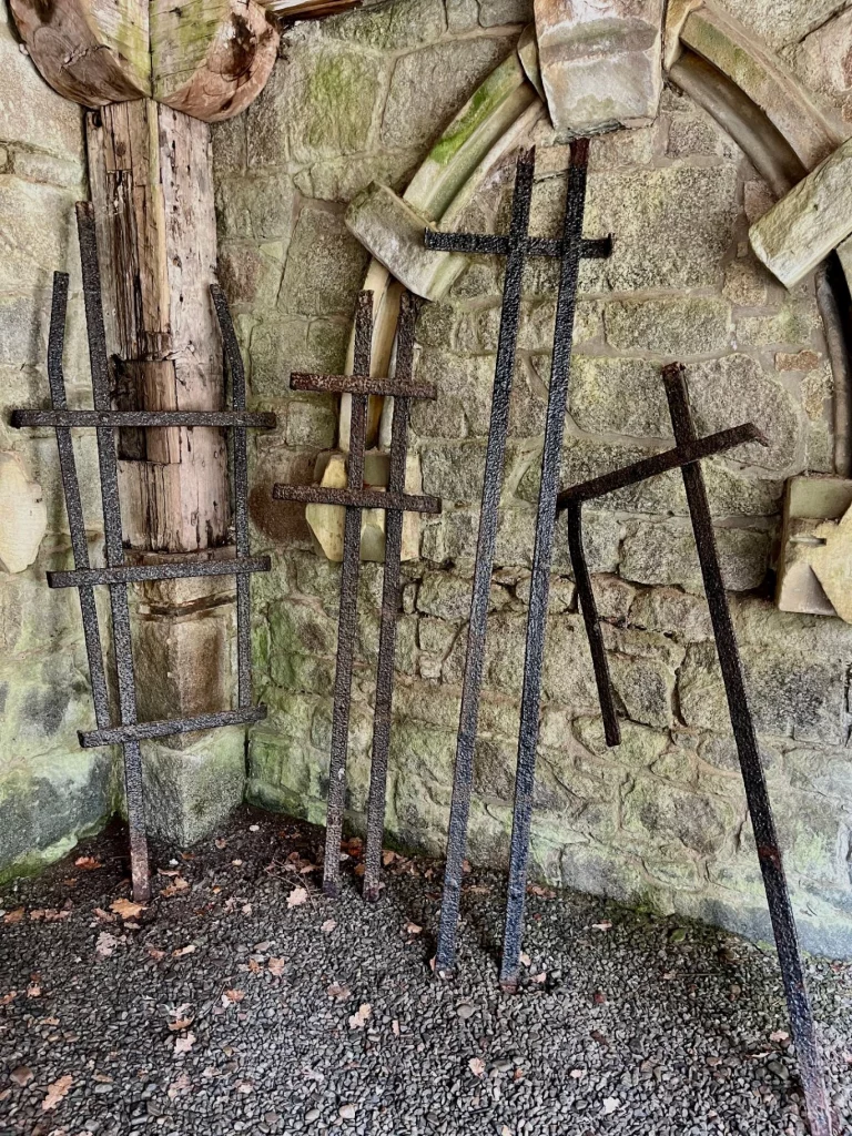 Nahaufnahme von drei stark verrosteten, kreuzförmigen Eisengittern, die gegen eine grobe Steinmauer im Außenbereich der Kirche lehnen. Im Hintergrund ist ein angedeuteter Rundbogen aus Stein zu sehen.