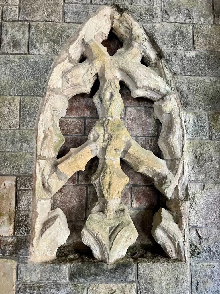 Ein freistehendes, spitzbogenförmiges Fenstergerüst aus hellem Stein, eingebaut in eine dunklere Bruchsteinmauer. Die filigranen Steinverstrebungen (Maßwerk) im Inneren bilden ein dekoratives Muster.