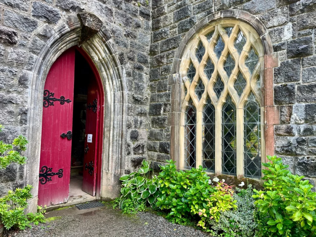Außenansicht der Kirche mit einer offenstehenden, zweiflügeligen Holztür in kräftigem Rot. Rechts daneben befindet sich ein großes Rundbogenfenster mit komplexem, rautenförmigem Steinmaßwerk, davor wachsen grüne Hostas.