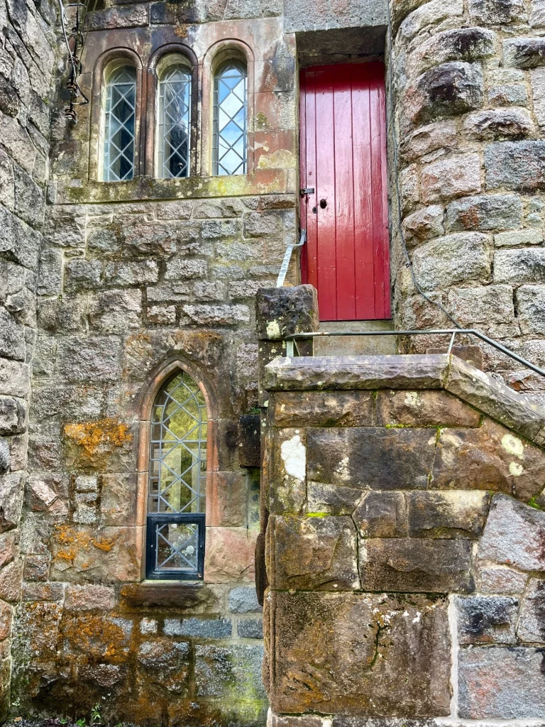 Außenansicht einer massiven Steinmauer aus unregelmäßigen Blöcken. Zu sehen sind ein spitzzulaufendes Fenster im Erdgeschoss, eine Dreiergruppe kleiner Rundbogenfenster darüber und eine schmale, leuchtend rote Tür, die über eine steinerne Außentreppe erreichbar ist. Flechten und Moose besiedeln die sonnenbeschienenen Steinflächen.