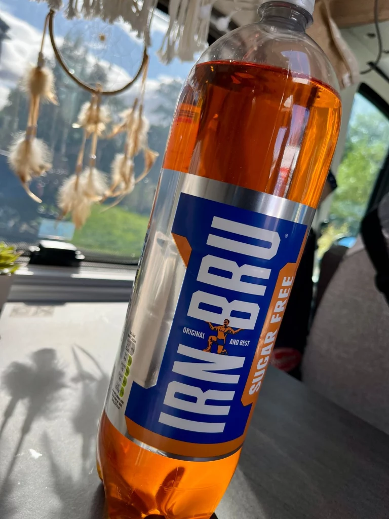 Nahaufnahme einer Kunststoffflasche mit dem orangefarbenen Erfrischungsgetränk Irn-Bru Sugar Free. Im Hintergrund ist ein helles Fenster mit einem Traumfänger zu sehen.
