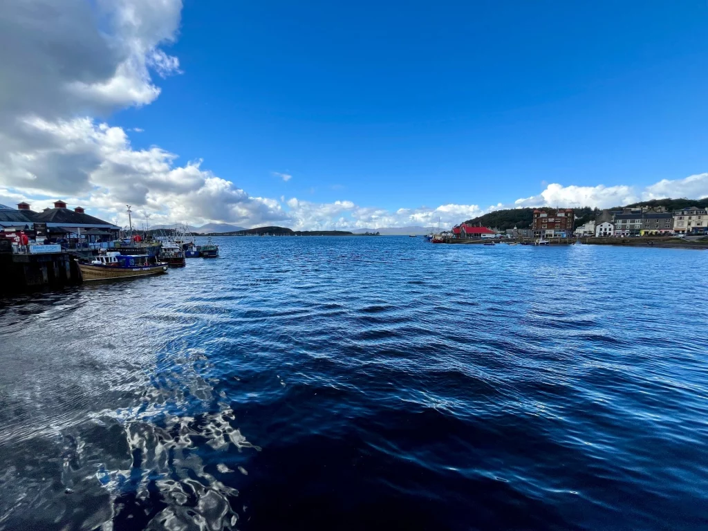Eine Weitwinkelaufnahme des Meeresarms in Oban. Im Vordergrund kräuselt sich dunkles, blaues Wasser. Am linken Rand sind Ausflugsboote an einem Kai vertäut, in der Ferne sind die grünen Silhouetten von Inseln und Bergen unter weißen Wolken zu sehen.
