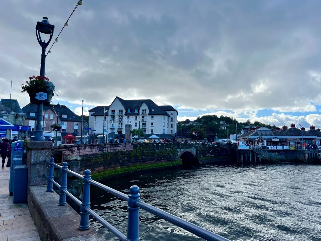 Ansicht einer alten, mit Moos und Algen bewachsenen Steinmauer am Meeresufer. Im Hintergrund steht ein großes weißes Apartmenthaus, rechts ist ein Teil eines Hafengebäudes mit rotem Dach zu sehen. Der Himmel ist bewölkt.