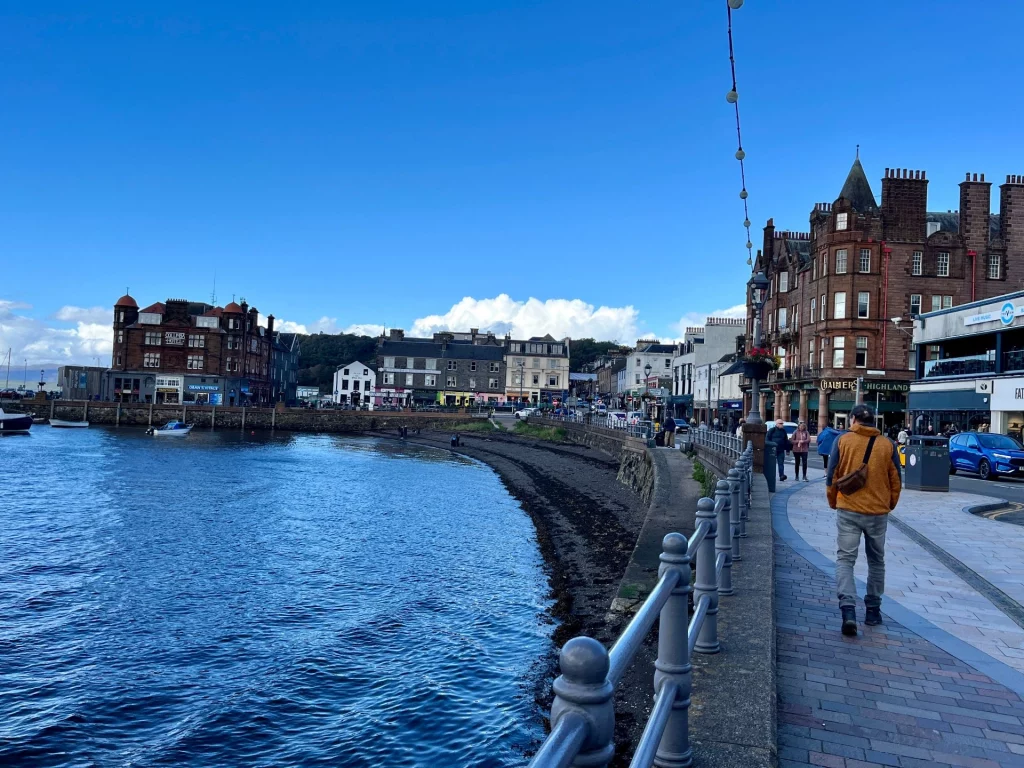 Blick von einer gepflasterten Uferpromenade mit Metallgeländer auf die Meeresbucht von Oban. Rechts befinden sich historische Gebäude aus rotem Sandstein, links das blaue Wasser und im Hintergrund bewaldete Hügel unter blauem Himmel.