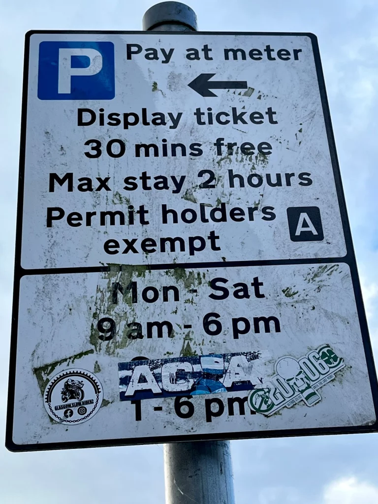 Ein weißes Verkehrsschild mit schwarzer Schrift erklärt die Parkregeln: "Pay at meter", "30 mins free" und "Max stay 2 hours". Das Schild ist teilweise mit Aufklebern, unter anderem von "Glasgow Slow Riders", beklebt.