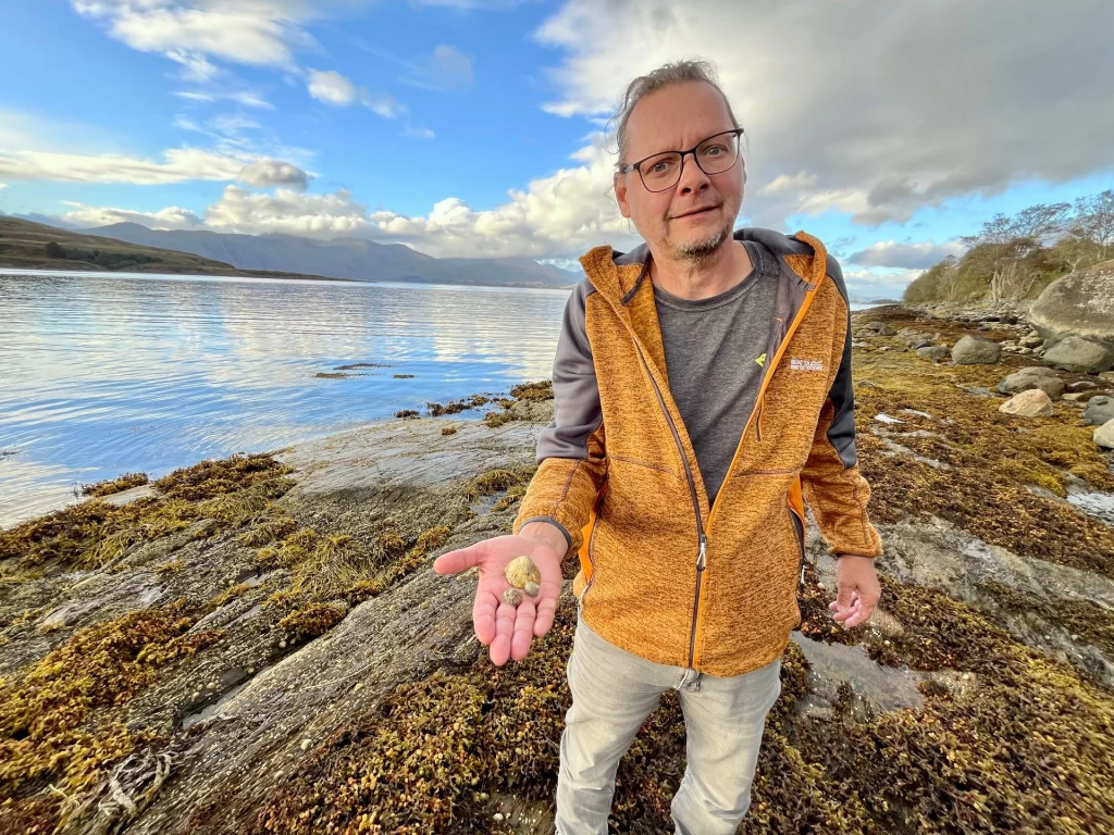 Ein Mann in einer gelben Outdoor-Jacke steht am Ufer und hält seine flache Hand in die Kamera, auf der drei kleine, verschiedene Muschelschalen liegen.