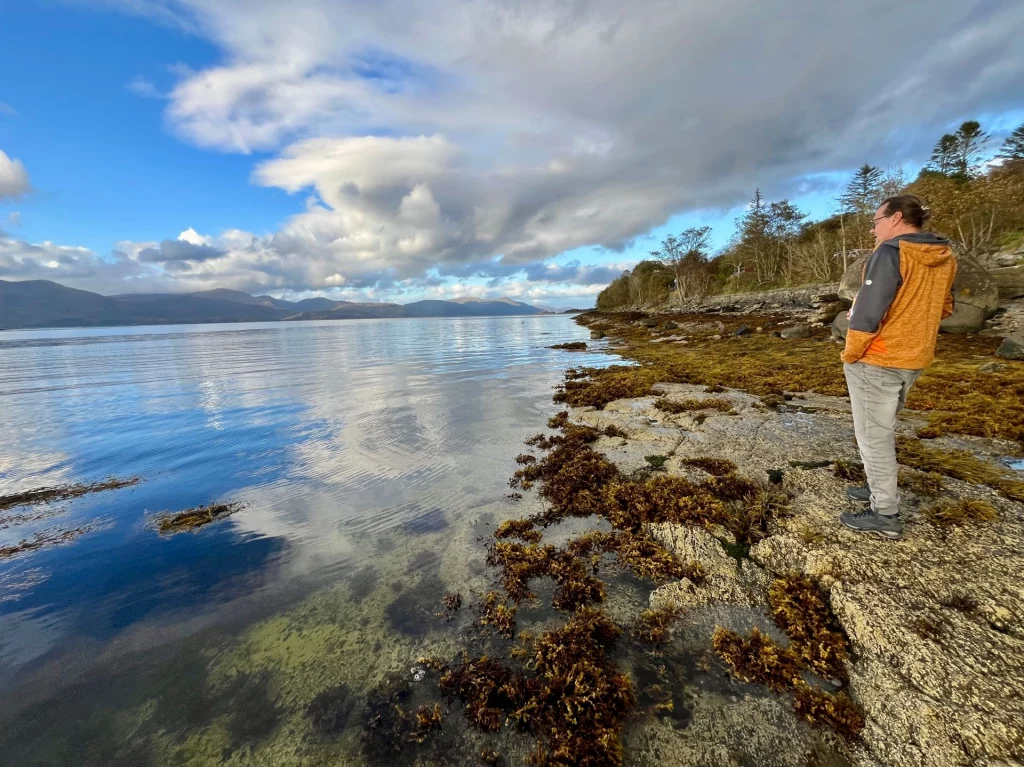 Ein Mann in einer orangen Jacke steht am felsigen, mit braunem Seetang bewachsenen Ufer eines weiten Sees und blickt auf das ruhige Wasser und die fernen Berge unter einem bewölkten Himmel.