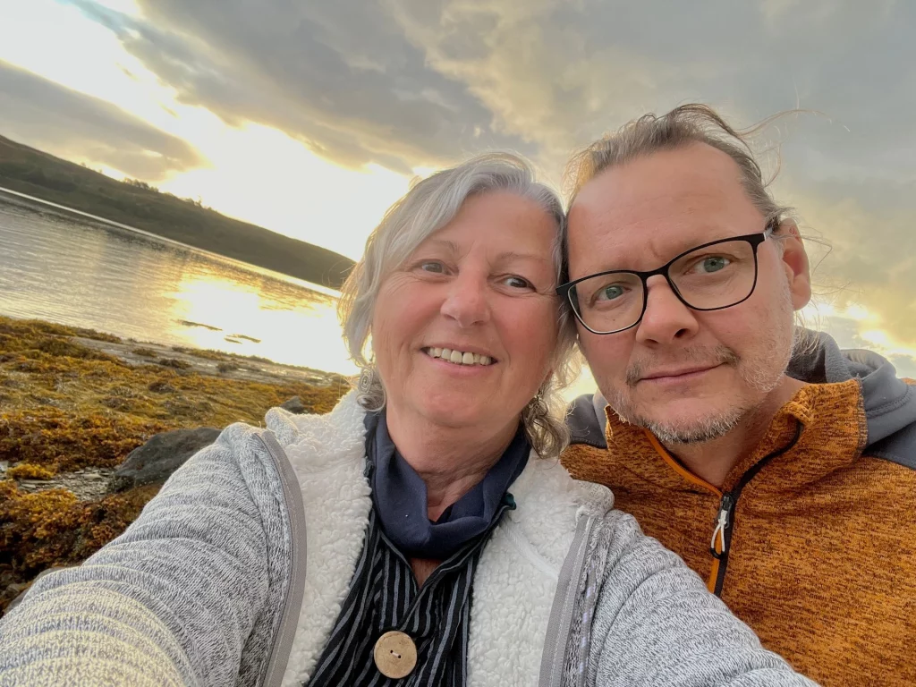 Ein lächelndes Paar macht ein Selfie vor einem See bei Sonnenuntergang. Im Hintergrund sind das Wasser und das dunkle, mit Seetang bedeckte Ufer zu sehen.