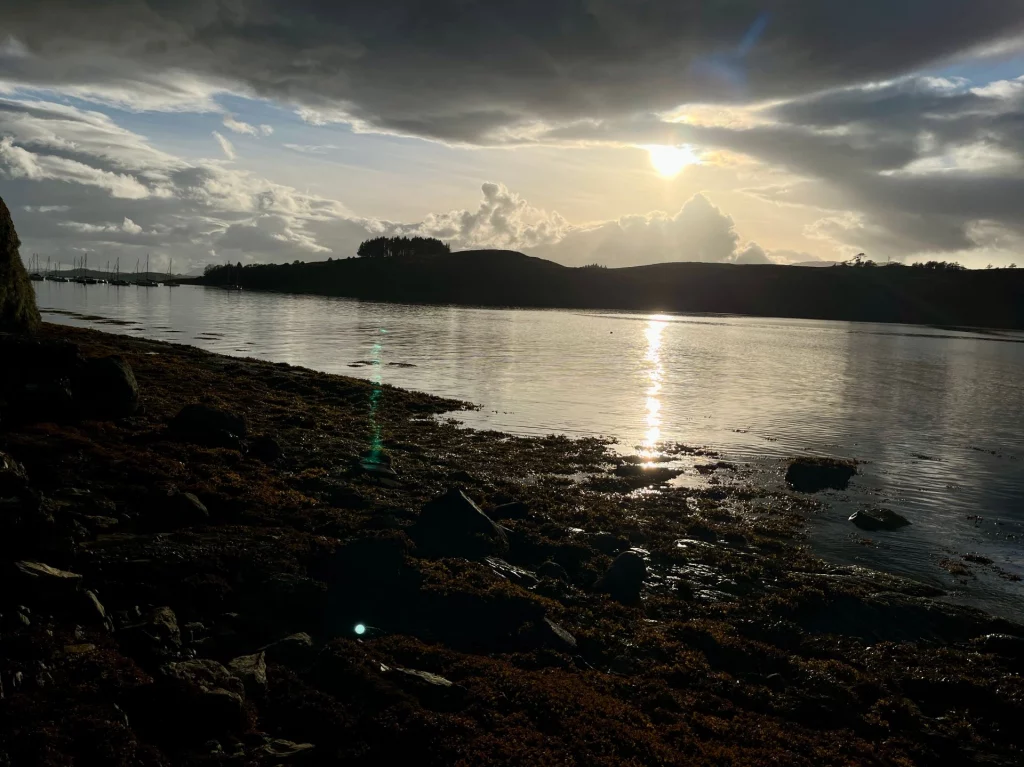 Landschaftsaufnahme gegen das Licht. Eine dunkle Uferlinie im Vordergrund kontrastiert mit dem glitzernden Wasser. Am Horizont sind dunkle Hügelketten unter einem weiten Himmel mit markanten Wolken zu sehen.