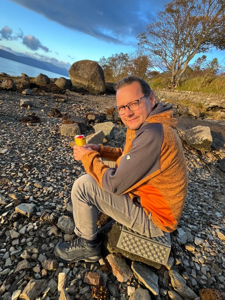 Ein Mann mit Brille und orange-grauer Outdoorjacke sitzt auf einem grauen Sitzkissen auf einem steinigen Strand. Er hält eine gelbe Getränkedose in den Händen und lächelt in die Kamera. Im Hintergrund sind das Meer und große Findlinge zu sehen.