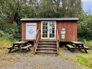 Eine rustikale Holzhütte mit der Aufschrift „Reception“ und dem Highland Titles Logo dient als Besucherzentrum. Davor stehen zwei hölzerne Picknicktische auf einem Kiesplatz, umgeben von grünem Gebüsch.