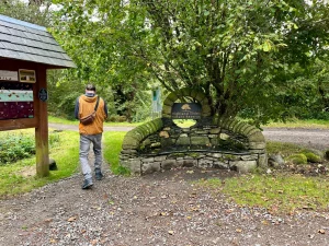Ein Mann in einer orangenen Jacke geht an einer hölzernen Informationstafel vorbei in Richtung eines dichten Waldstücks. Neben dem Weg steht eine kunstvoll gestaltete Steinbank unter einem großen Baum.