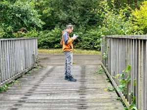 Ein Mann steht im Profil auf einer stabilen Holzbrücke und schaut auf sein Smartphone, vermutlich um die Koordinaten zu prüfen. Die Brücke führt über einen hellen Untergrund in einen bewaldeten Bereich.