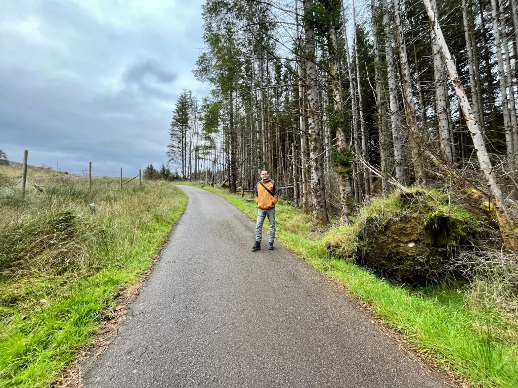 Eine asphaltierte Straße schlängelt sich durch eine hügelige Landschaft; am Rand steht eine Person in einer orangenen Jacke vor hohen Nadelbäumen