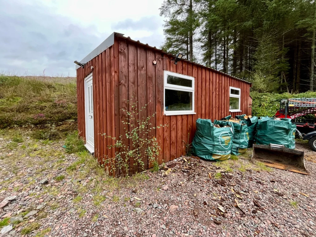 Eine längliche, rustikale Holzhütte mit weißen Fensterrahmen steht auf einem Schotterplatz neben gestapelten Säcken.