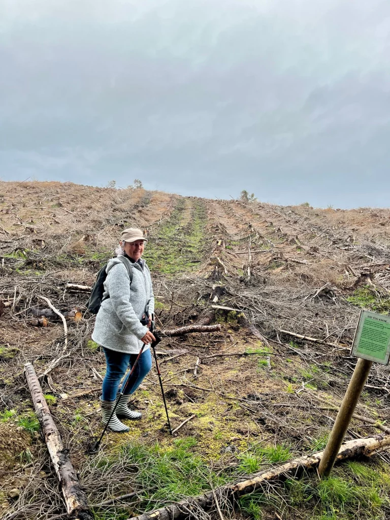 Karin steht lächelnd mit Nordic-Walking-Stöcken auf einem Baumstumpf inmitten einer großen Rodungsfläche. Vor ihr im Boden steckt ein kleines grünes Schild mit der Aufschrift „Highland Titles – Your Plot“. Im Hintergrund ist eine weite Fläche mit vielen Baumstümpfen und ein ferner Nadelwald zu sehen.