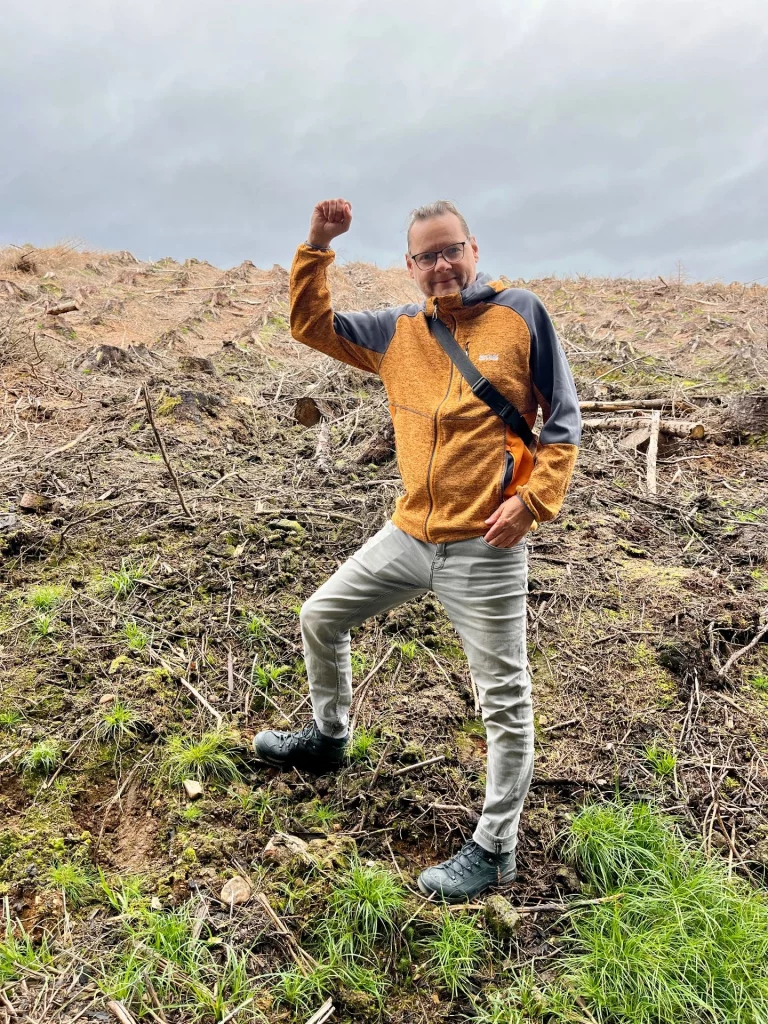 Ingo steht triumphierend mit einer geballten Faust auf demselben Baumstumpf hinter dem „Highland Titles“-Schild. Er trägt eine orangefarbene Outdoorjacke und eine Umhängetasche. Die Szenerie zeigt die weite, zur Wiederaufforstung bereite Fläche unter einem bewölkten Himmel.