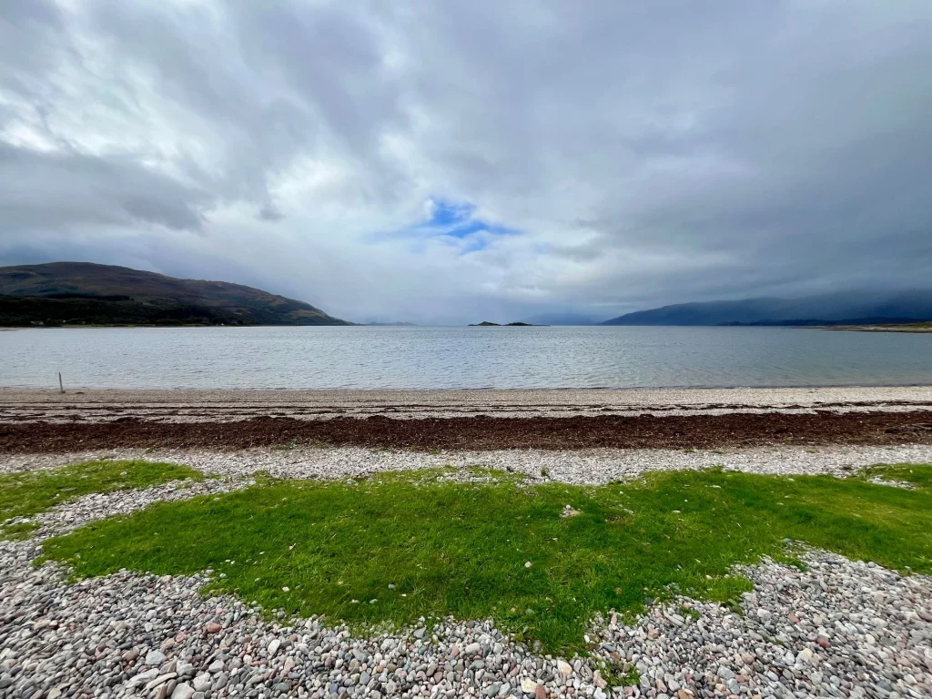 Eine Weitwinkelaufnahme eines steinigen Strandes mit einem Streifen aus dunklem Seegras. Im Vordergrund wächst leuchtend grünes Gras zwischen grauen Kieselsteinen. Das Wasser ist ruhig, und am Horizont sind dunkle Berge unter einer dichten, grauen Wolkendecke mit einer kleinen blauen Lücke zu sehen.