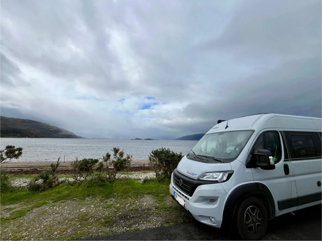Ein hellgrauer Kastenwagen-Camper steht am rechten Bildrand geparkt auf einem Schotterplatz. Links daneben öffnet sich der Blick auf eine weite Wasserfläche und die gegenüberliegende Hügelkette unter einem bewölkten Himmel. Ein paar grüne Büsche trennen den Parkplatz vom Strandbereich.