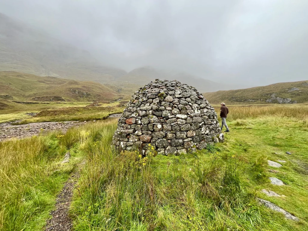 Ein kuppelförmiger Steinhaufen (Cairn) steht auf einer grünen Wiese neben einem schmalen Wanderpfad. Im Hintergrund erstrecken sich sanfte, grasbewachsene Hügel unter einem wolkenverhangenen, grauen Himmel. Eine Person steht rechts im Hintergrund.
