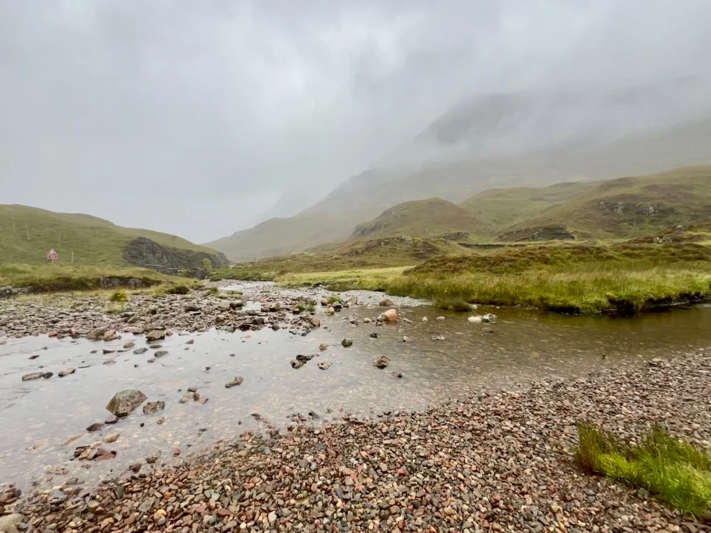 Ein flacher, klarer Bachlauf mit Kieselsteinen im Vordergrund und nebeligen Hügeln im Hintergrund.