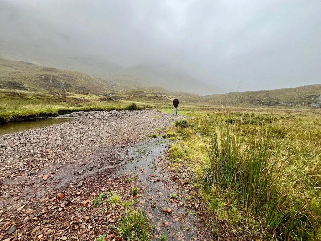 Eine Weitwinkelaufnahme eines flachen, steinigen Flussufers in einer weiten Tallandschaft. Ein Wanderer ist in der Ferne zu sehen. Die Vegetation besteht aus hohem Gras und Heidekraut, während die Berggipfel vollständig im Nebel verschwinden.