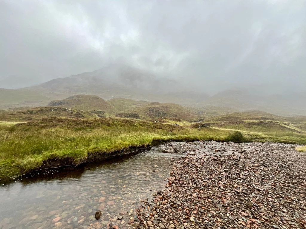 Ein schmaler Fluss, der sich durch die typische Highland-Vegetation in einem weiten Tal zieht.